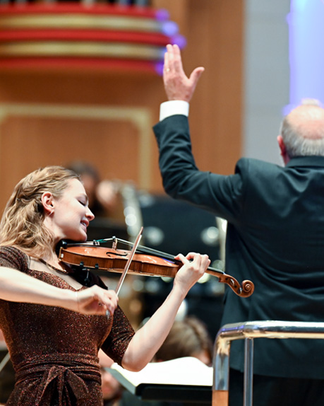 A female violinist performing in front of a conductor, conducting an orchestra behind her.